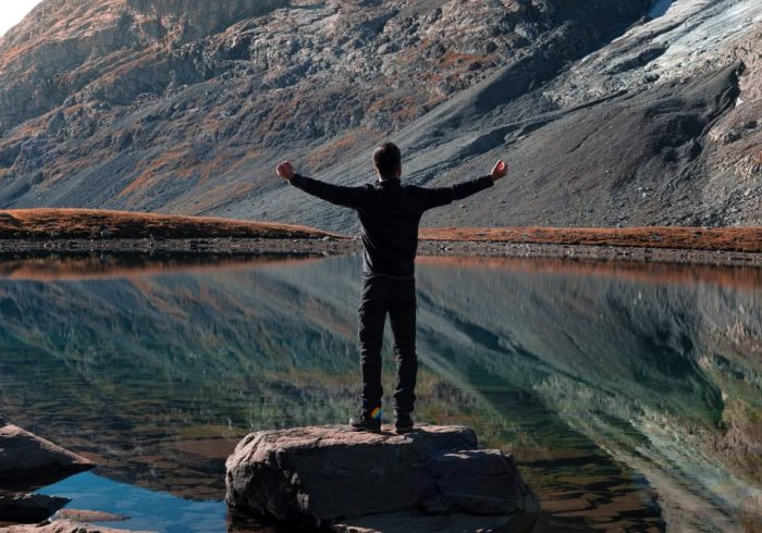 A man stands with outstretched arms by a mountain lake, reflecting rocky slopes in Auvergne-Rhône-Alpes.